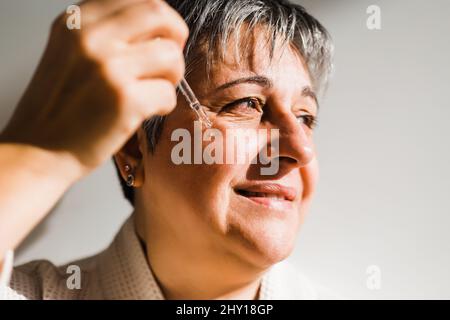Side view of smiling elderly Woman using moisturizing serum while having skincare procedure and looking away Stock Photo