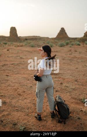 Full body back view of female tourist with binoculars looking into distance while standing in desert area with rocky formations Stock Photo
