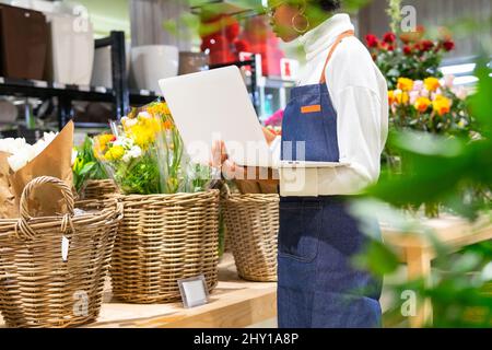 Side view of African American female in apron checking various colorful flowers while working on laptop in light floral shop Stock Photo