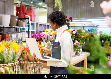Side view of African American female in apron checking various colorful flowers while working on laptop in light floral shop Stock Photo