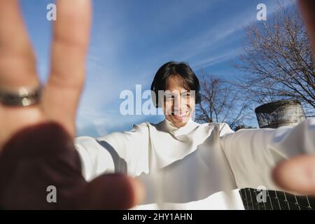 Positive Indian male taking self portrait and looking away while listening to music in wireless headphones on sunny street in city Stock Photo