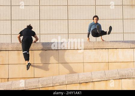Full body male athletes in sportswear and sneakers parkour jumping above stone roof border while exercising on sunny day on street Stock Photo