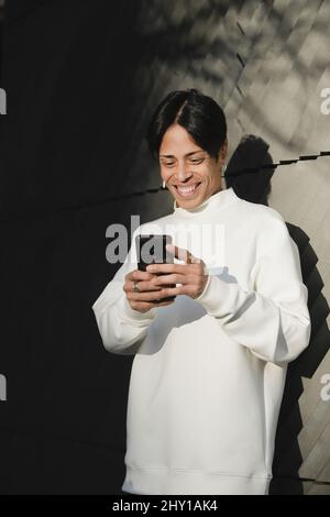Smiling Asian man in white jumper standing on street concrete wall and reading messages on smartphone Stock Photo