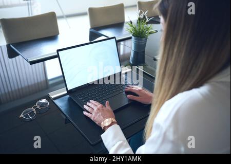 Side view of crop anonymous female employee browsing netbook while working at table with eyeglasses in light modern office during workday Stock Photo