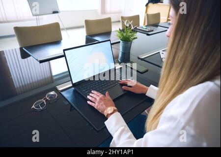 Side view of crop anonymous female employee browsing netbook while working at table with eyeglasses in light modern office during workday Stock Photo