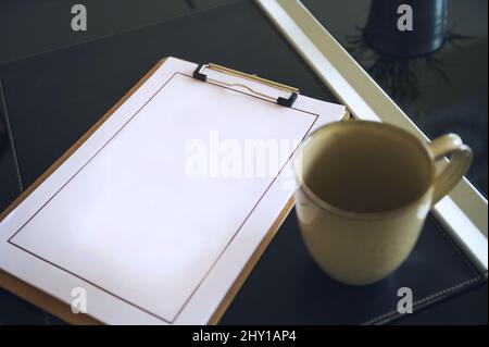From above of clipboard with empty white sheet placed on desktop with ceramic cup in light modern office of company Stock Photo
