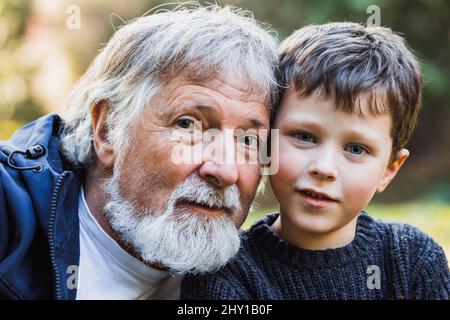 Positive aged man with gray beard and hair and cute smiling kid with blue eyes looking at camera in forest against blurred background Stock Photo