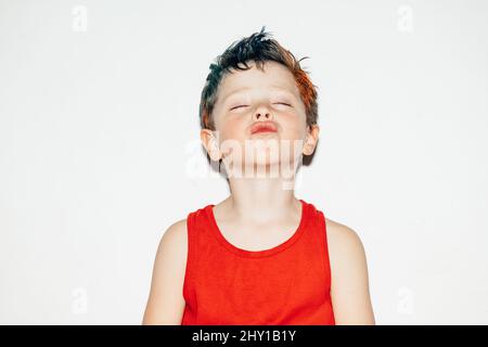 Naughty boy in t shirt with colorful dyed hair pouting lips with closed eyes while standing on white background in light studio Stock Photo