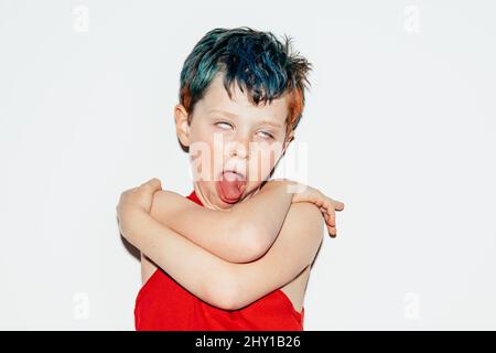 Mischievous boy with colorful dyed hair rolling eyes and showing tongue on white background in light room Stock Photo