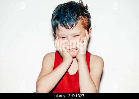 Rebellious boy with dyed hair looking away and touching face with hands while standing on white background in room Stock Photo