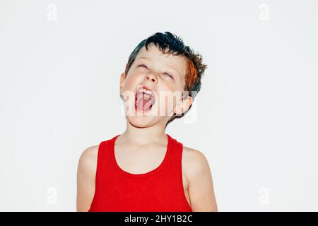 Mischievous boy with colorful dyed hair rolling eyes and showing tongue on white background in light room Stock Photo