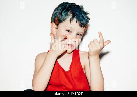 Mischievous boy with colorful dyed hair showing gesture with fingers on white background in light room Stock Photo