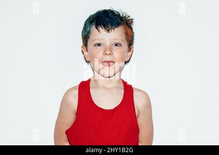 Mischievous boy with colorful dyed hair grinning and showing tongue while looking at camera on white background in light room Stock Photo