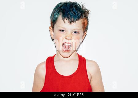 Mischievous boy with colorful dyed hair grinning and showing teeth while looking at camera on white background in light room Stock Photo