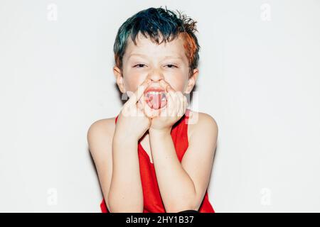 Mischievous boy with colorful dyed hair showing tongue and touching nose on white background in light room Stock Photo