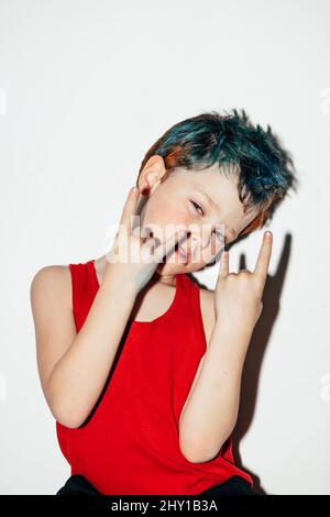 Mischievous boy with colorful dyed hair showing rock and roll gesture on white background in light room Stock Photo