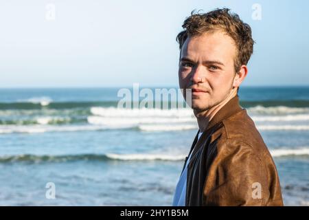 Young male in leather jacket looking at camera while standing on coast near waving sea against blue sky in coastal area Stock Photo