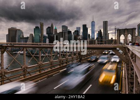 Long exposure of vehicles driving on road on suspension Brooklyn Bridge connecting East River shores in New York City against cloudy gray sky Stock Photo