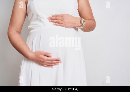 Crop anonymous pregnant female with wristwatch in elegant white dress touching belly while standing on white background in light studio Stock Photo