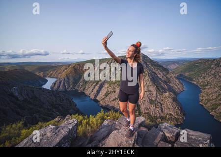 From above of full lenght adult female tourist in sportswear standing on top and taking selfie on smartphone while enjoying picturesque landscape Stock Photo