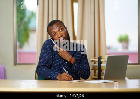 Young Black guy from Ghana working on his desk surprised with some news talking on the phone Stock Photo