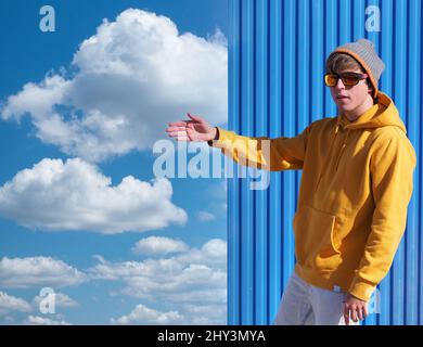 Cheerful young man in a yellow sweatshirt and wool hat pointing to the blue sky with clouds Stock Photo