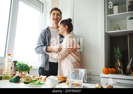 Smiling man hugging caucasian woman standing in the kitchen during the breakfast preparation process. Cooking, together, kitchen, relationship Stock Photo