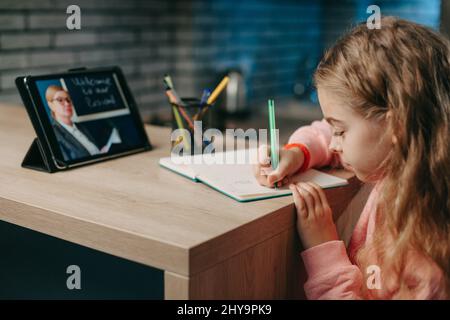 Schoolgirl talking with remote teacher tutor on video conference, online virtual class, while writing in her notebook. Coronavirus pandemic. Kid learn Stock Photo