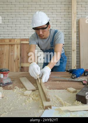 Asian carpenter with dustproof glasses drawing a line on a piece of wood, preparing to cut into sections. Morning work atmosphere in the workshop room Stock Photo