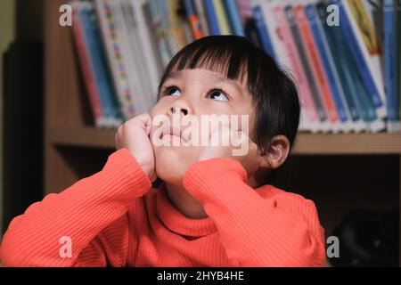 Portrait of cute child looking up and thinking with hands on chin in living room. school child thinking Stock Photo