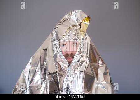 Lost hiker wrapped in an emergency survival blanket, isolated on blue wall. Stock Photo