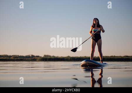 Low-angle of Caucasian woman standing on sup board with one oar in hands looking up at sky on crystal blue lake wearing summer swimming dress. Active Stock Photo