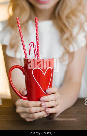 A red mug with a heart pattern with cocoa and paper tubes in the hands of a woman. Valentine's Day Concept Stock Photo