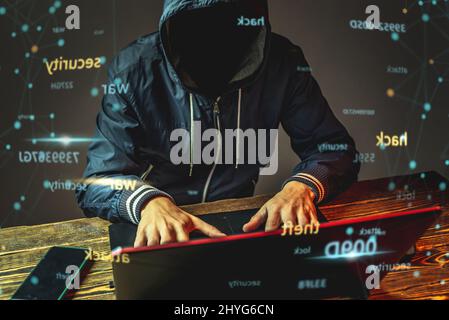 A hacker anonymous in a hood is typing on a laptop keyboard in a dark room. The concept of cyber warfare and Dos attacks Stock Photo