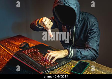 A hacker anonymous in a hood is typing on a laptop keyboard in a dark room. The concept of cybercrime fraud and identity theft Stock Photo