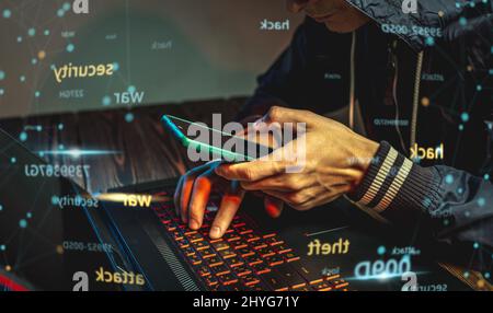 A hacker with a phone is typing with his hands on a laptop keyboard in a dark room. The concept of cyber warfare and Dos attacks Stock Photo