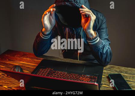 A hacker anonymous in a hood is typing on a laptop keyboard in a dark room. The concept of cyber warfare and Dos attacks Stock Photo