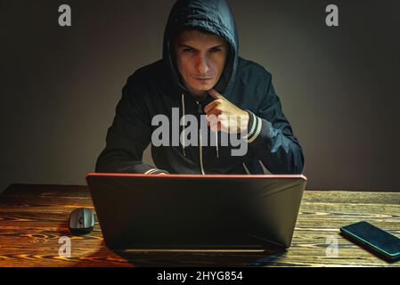 A hacker in a hood with a phone is typing on a laptop keyboard in a dark room. The concept of cyber warfare and Dos attacks Stock Photo