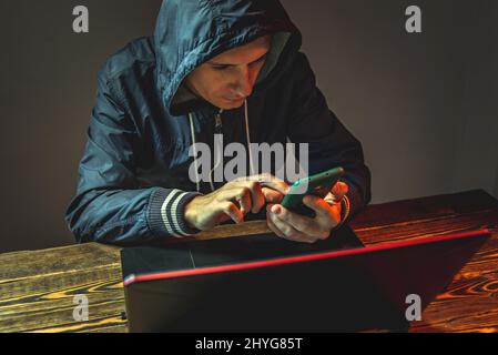 A hacker in a hood with a phone is typing on a laptop keyboard in a dark room. The concept of cyber warfare and Dos attacks Stock Photo