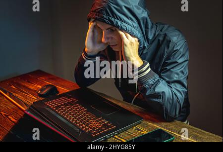 A hacker in a hood with a phone is typing on a laptop keyboard in a dark room. The concept of cybercrime fraud and identity theft Stock Photo