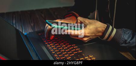 A hacker with a phone is typing with his hands on a laptop keyboard in a dark room. The concept of cybercrime fraud and identity theft Stock Photo