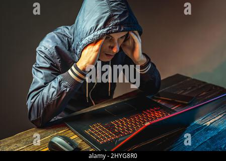 A hacker in a hood with a phone is typing on a laptop keyboard in a dark room. The concept of cyber warfare and Dos attacks Stock Photo