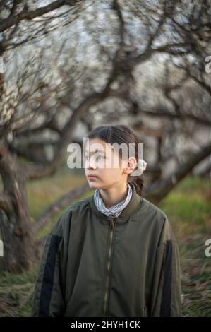 Profile portrait of a happy pre teen girl in a park. Child wearing a green jacket and turtleneck. Blooming trees in the background. Springtime. Stock Photo