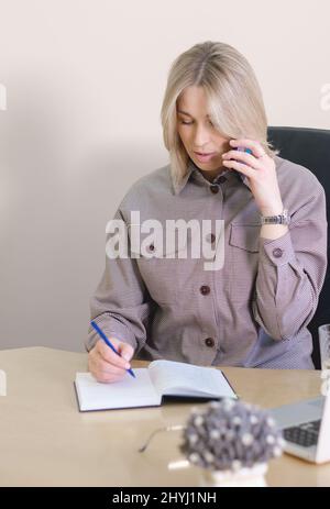 woman freelancer speaking to client by phone and making notes in a notebook. vertical shot of blond working woman writing notes Stock Photo