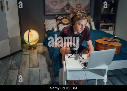 Home sound studio young teenager portrait using laptop computer and Headphones, playing guitar and recording voice music with microphone in the kid's Stock Photo