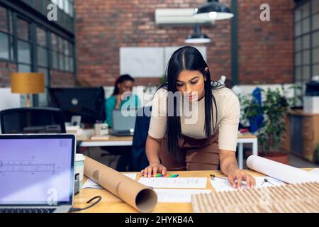 Biracial young female architect working over blueprint by building model at desk in office Stock Photo
