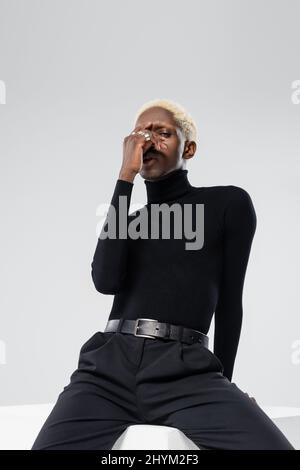 young african american man in turtleneck touching face while sitting on white cube isolated on grey Stock Photo
