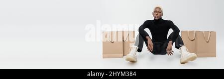 full length of blonde african american man sitting near paper bags on grey, banner Stock Photo