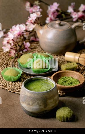 Asian rice dessert sweet green matcha mochi with cup of frothed matcha tea and green matcha powder in ceramic plate on brown table. Teapot and spring Stock Photo