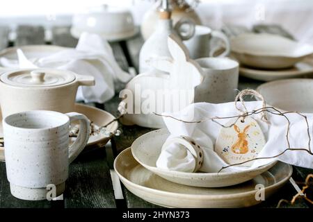 Rustic Easter table setting with empty craft ceramic tableware, plates and bowls, Easter bunny rabbit decorations, vase on wooden table in front of th Stock Photo